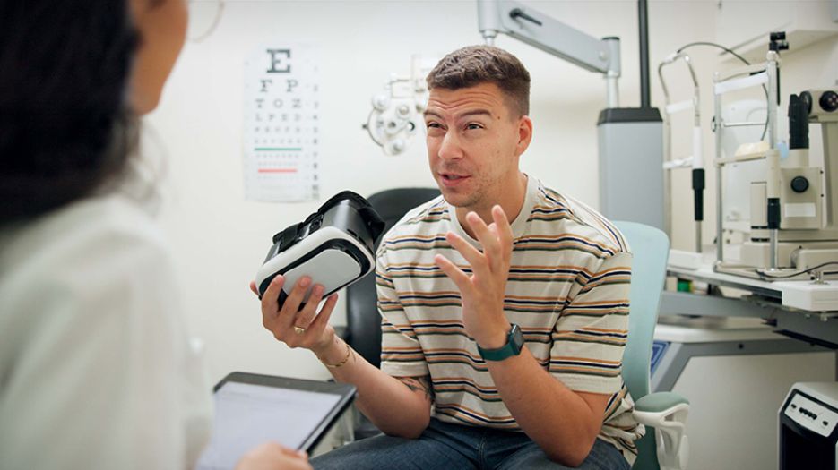 White man in striped shirt sitting in optometrist's office, holding a VR headset, talking to female doctor who is partially out of frame